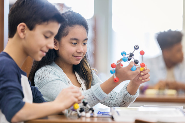 Smiling schoolgirl looks at a model of the molecular structure while working on a science assignment with her friend